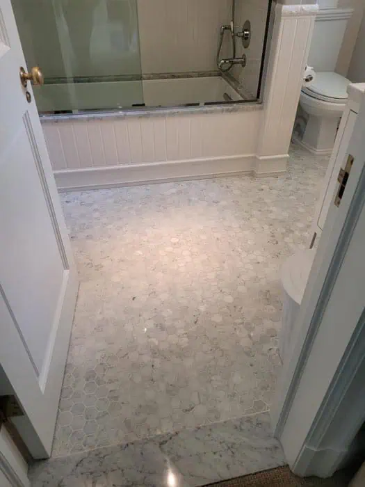 A bathroom with hexagonal marble tile flooring, showcasing the elegance of stone restoration Manhattan NY. It features a glass-enclosed bathtub with shower fixtures and a white toilet. The walls and door are also white, creating a clean, minimalist look.