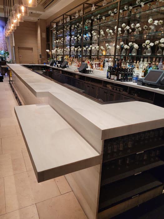 A modern bar interior featuring a long white countertop with clean lines, reminiscent of expert marble restoration in Astoria, NY. Behind the bar are shelves filled with bottles and decorative items, including vases with flowers. Warm lighting creates an inviting atmosphere.