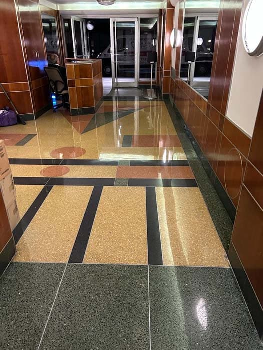 A shiny, modern lobby with geometric-patterned terrazzo flooring in shades of green, brown, and beige. The wood-paneled walls gleam like they've had expert stone restoration, perhaps from Manhattan NY. A reception desk sits in the backdrop near a windowed entrance.