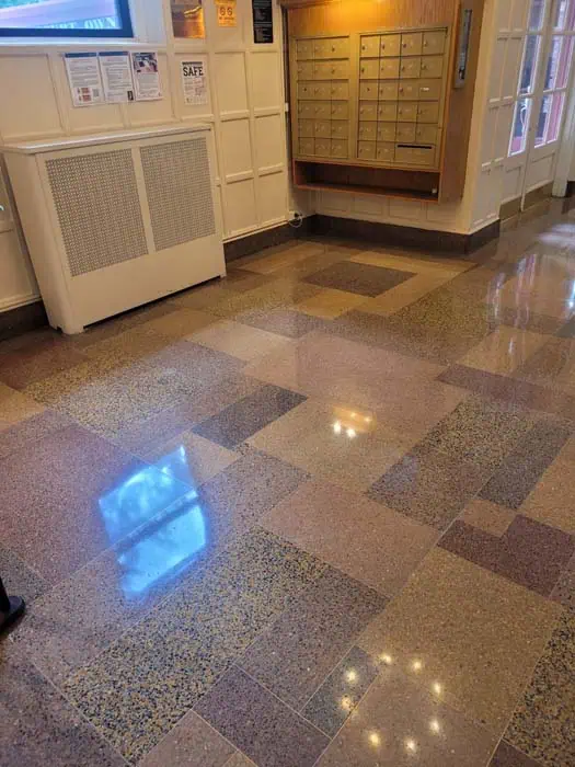 A polished hallway with a geometric-patterned terrazzo floor suggests recent stone restoration in Manhattan, NY. On the right, a set of mailboxes; to the left, a wall-mounted radiator and small notice board. A window above reflects light on the shiny floor.