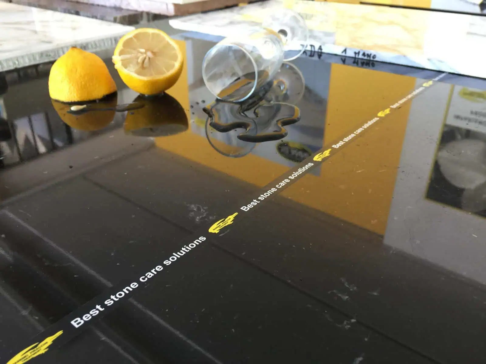 A polished black stone countertop with a sliced lemon and an overturned glass spilling liquid. A strip with the text "Best stone care solutions" runs across the surface.