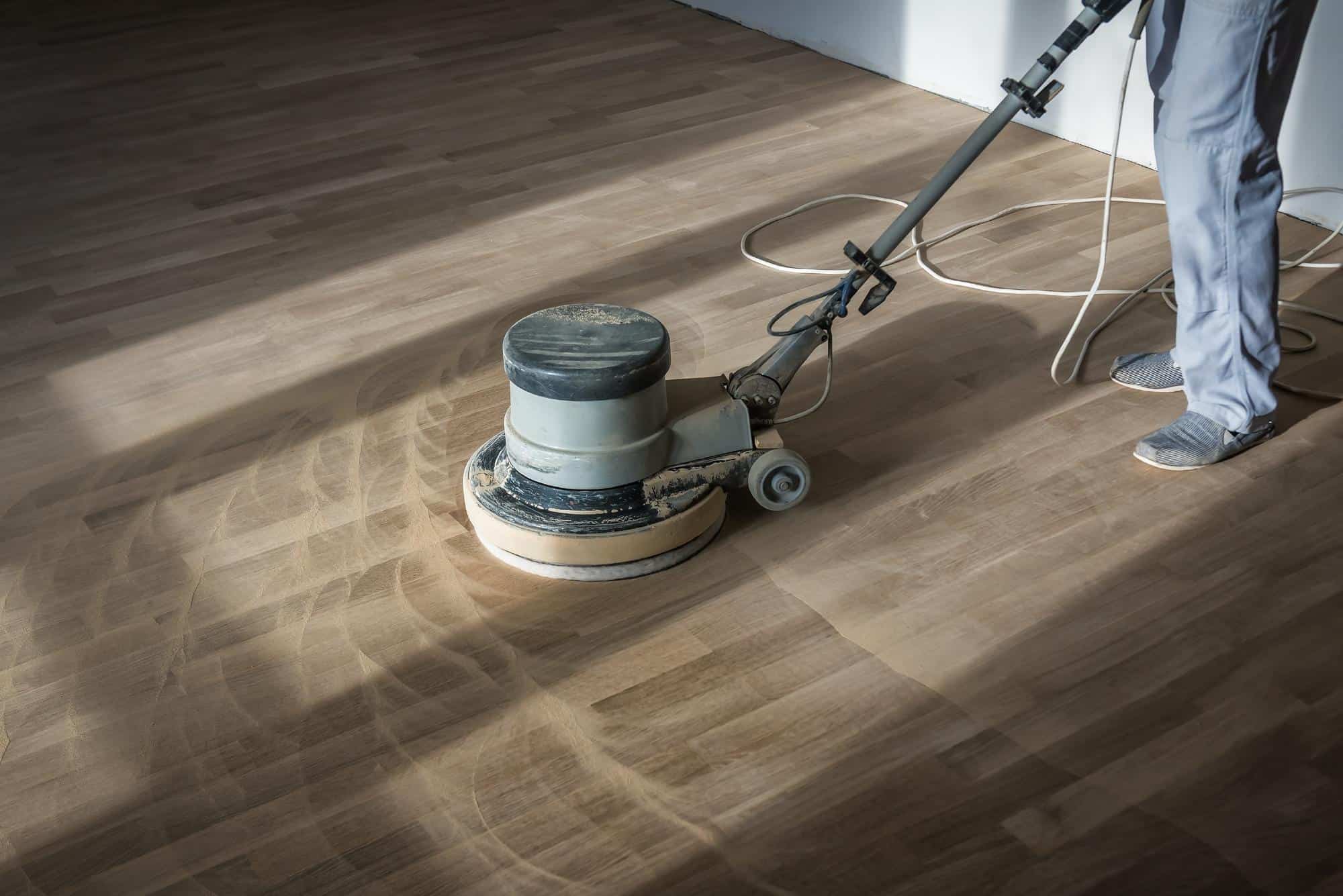 A person uses a floor polishing machine on a wooden floor, leaving circular cleaning marks on the surface. Only the person's lower legs are visible, along with the electrical cord trailing behind.