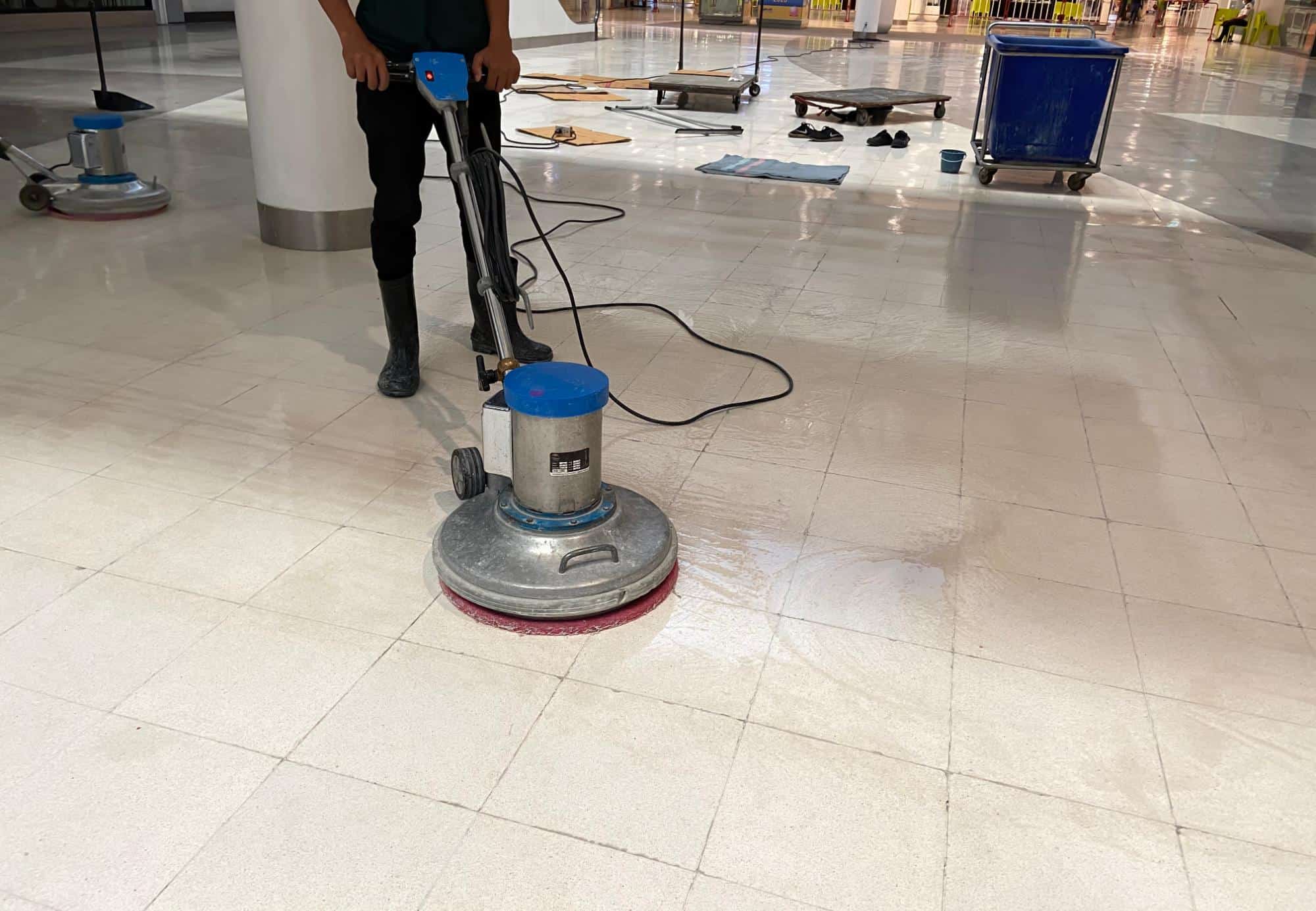 A person is using a floor polishing machine to clean shiny white tiles in a large indoor space. Nearby are cleaning supplies, a blue trolley, and some equipment on the floor.