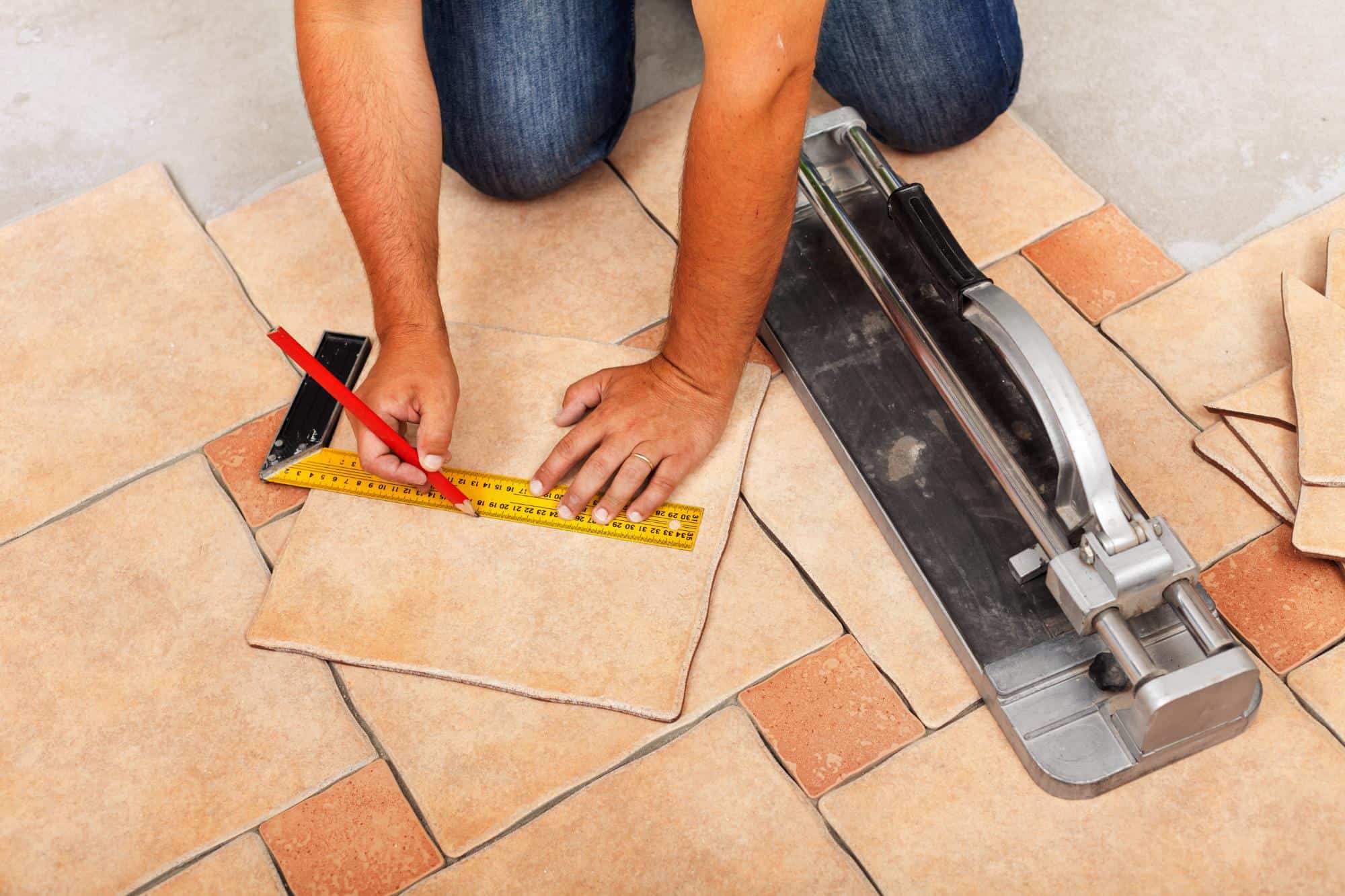 A person uses a pencil and tape measure to mark a ceramic tile for cutting, surrounded by tiles on the floor, with a tile cutter nearby.