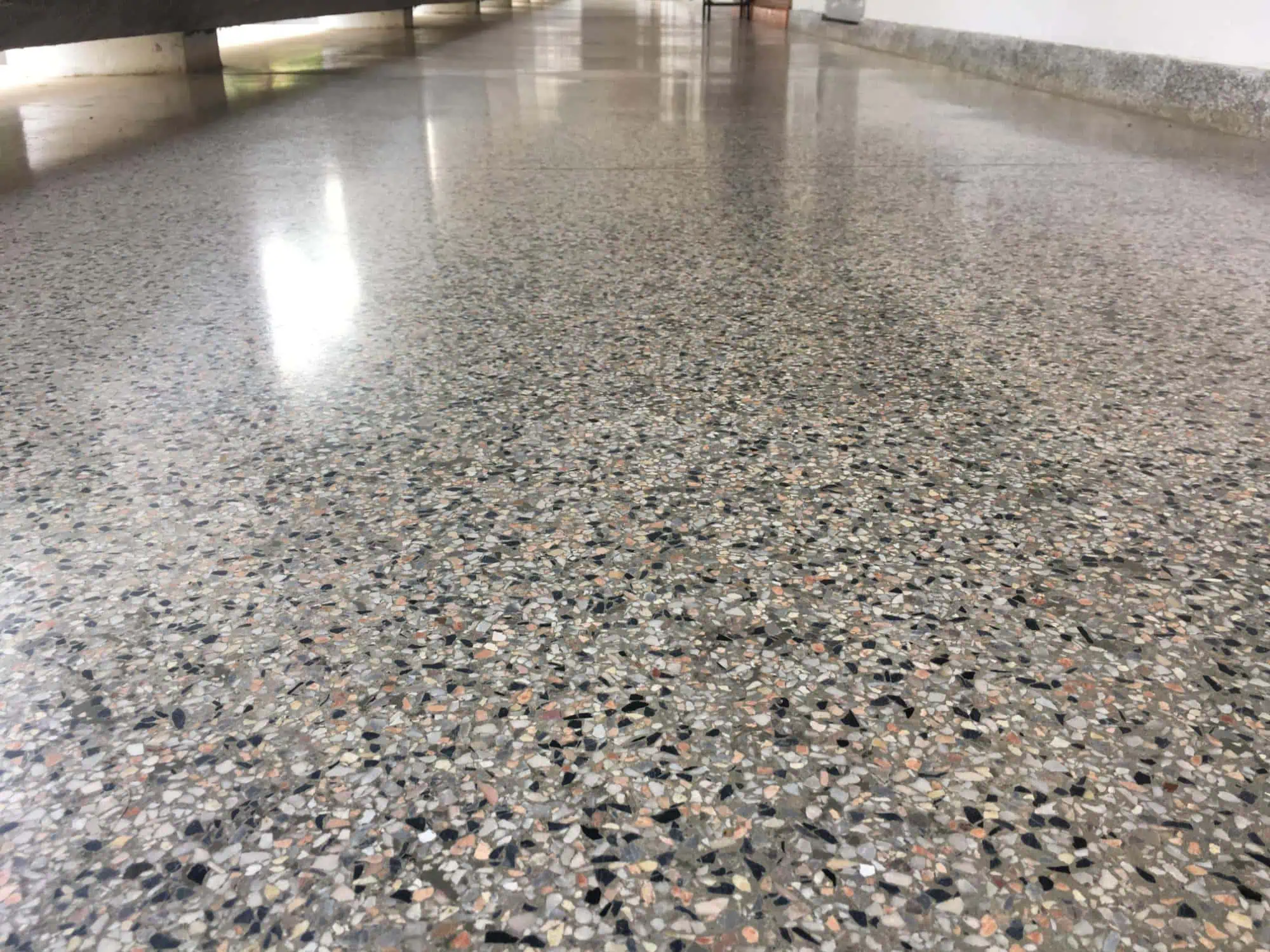 A close-up view of a polished terrazzo floor in a hallway, showing a mixture of small, multicolored stone chips embedded in the smooth surface. The corridor extends into the distance with walls on either side.