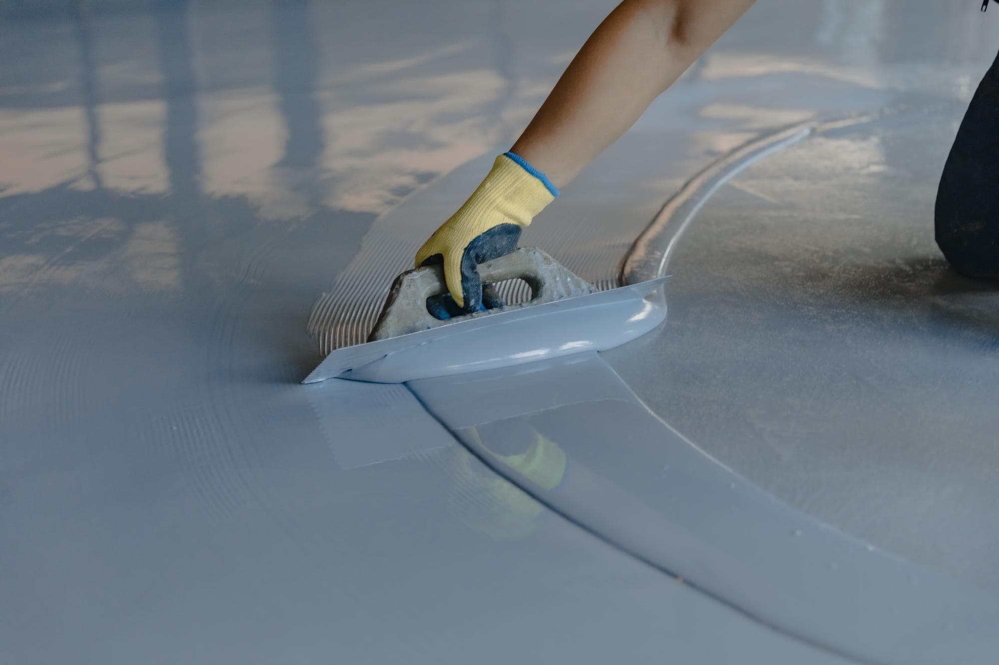 A person wearing a yellow glove uses a trowel to spread a layer of gray epoxy or concrete flooring, creating a smooth, glossy surface.