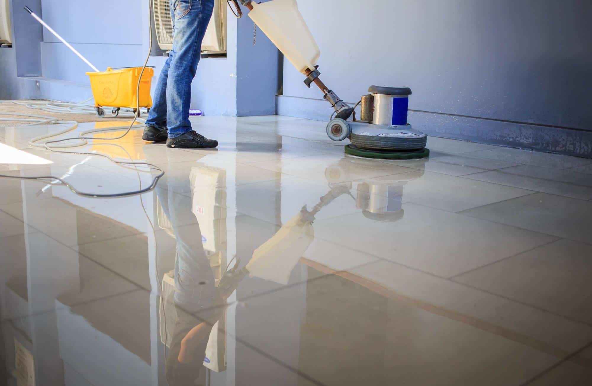 A person uses a floor polishing machine on shiny, tiled flooring, with a yellow mop bucket and cleaning tools visible in the background. The floor reflects the worker and equipment.