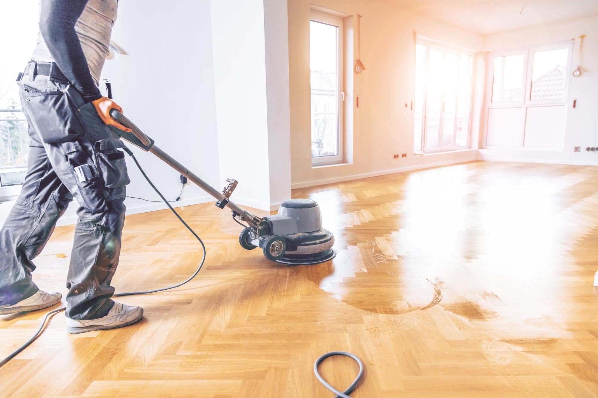 A person using a floor polishing machine on a wooden parquet floor in a bright, empty room with large windows and sunlight streaming in.