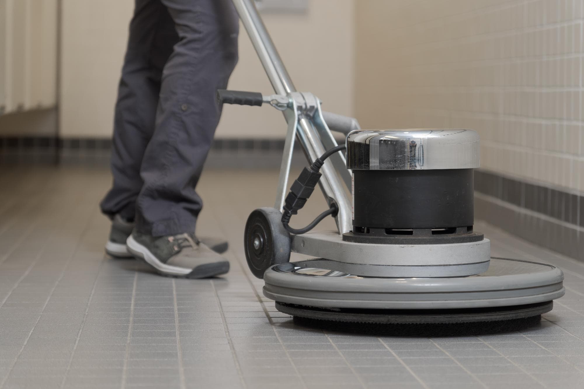 A person operating a floor cleaning machine on a tiled floor in a hallway, wearing gray pants and sneakers. The image focuses on the lower half of the person and the cleaning equipment.