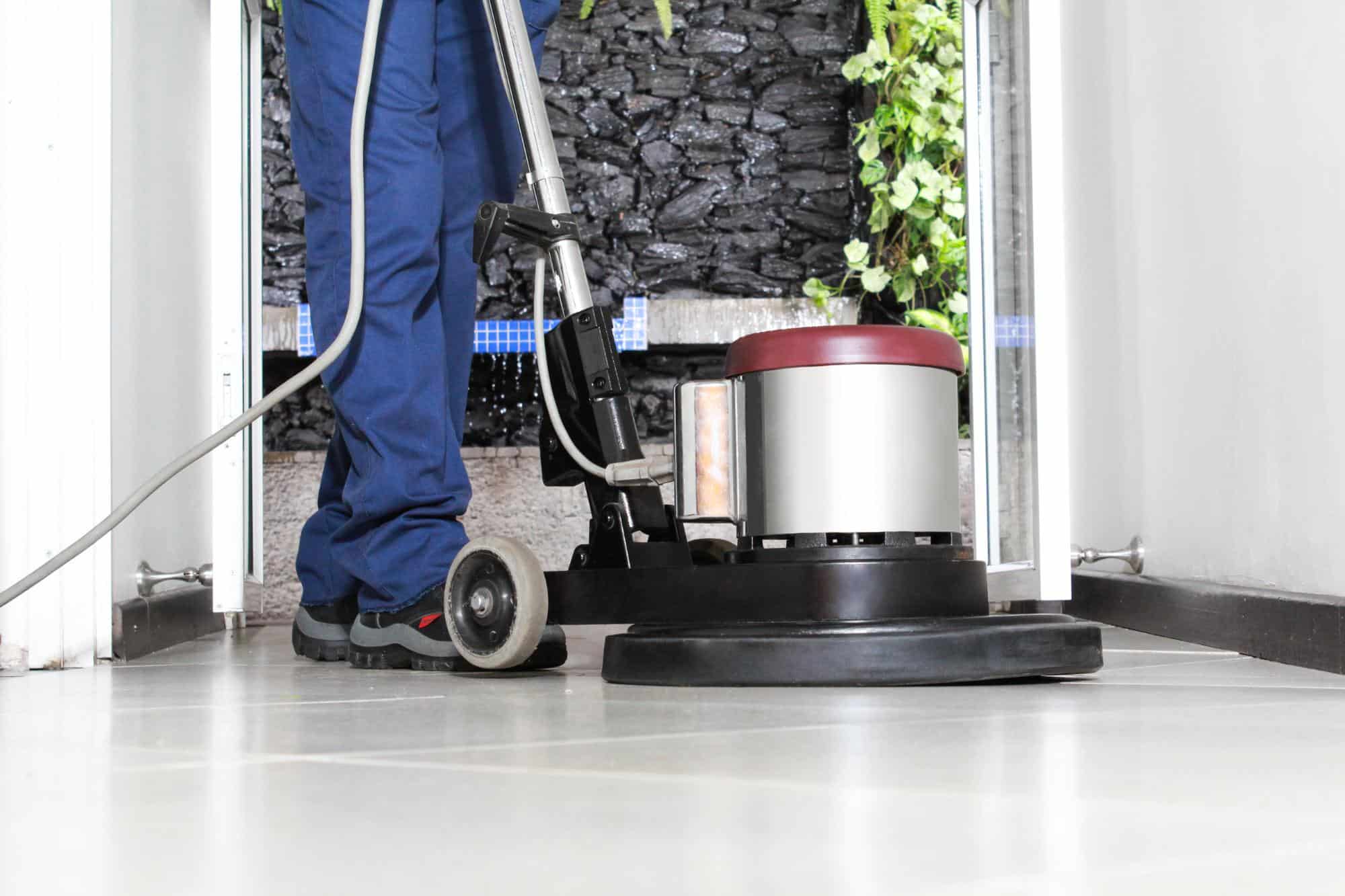 A person in blue pants and work shoes uses a floor polishing machine to clean shiny tile flooring in a bright hallway with an open door and plants outside.