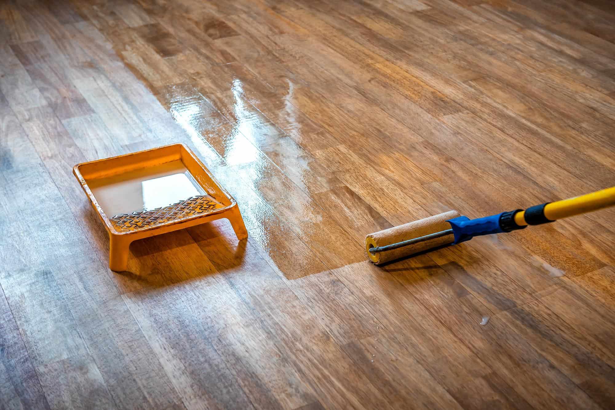 A close-up of a wooden floor being varnished with a roller brush, beside an orange paint tray filled with varnish. The section being coated appears shinier than the unfinished area.