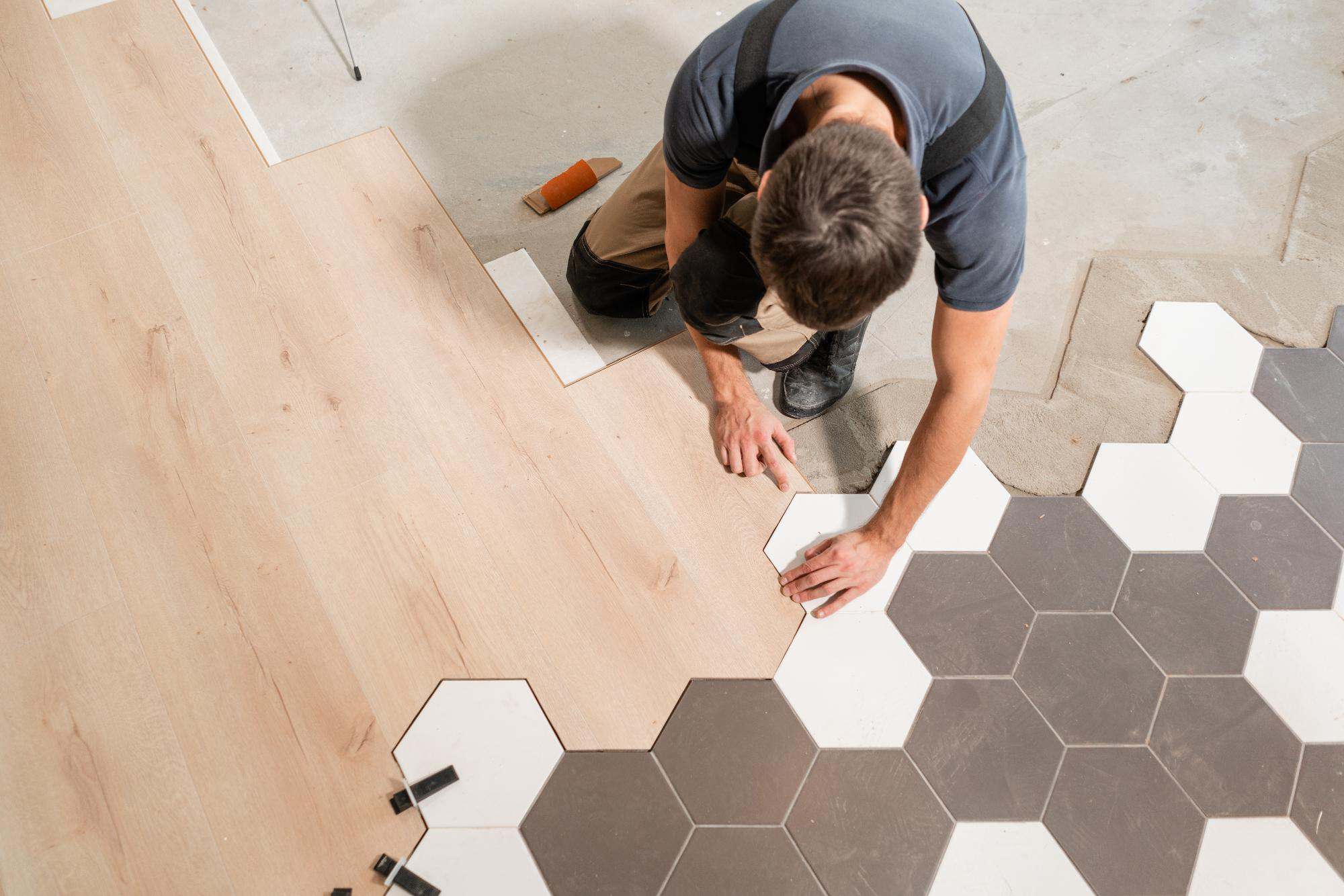 A person kneels on the floor, installing hexagonal tiles next to a section of wooden laminate flooring, carefully aligning the tiles where the two surfaces meet.