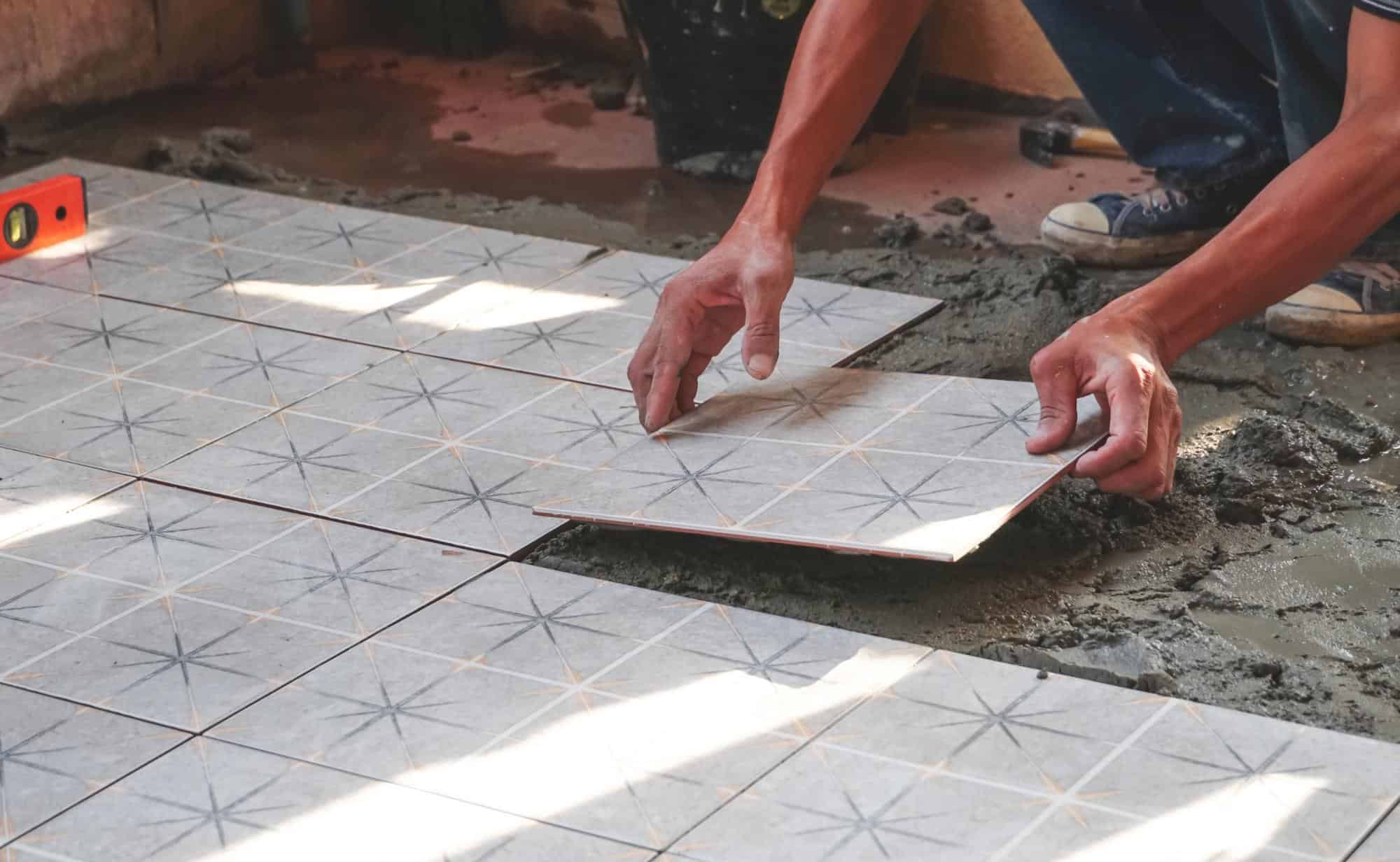 A person carefully placing a ceramic floor tile onto wet mortar, aligning it with other tiles during a flooring installation project. Sunlight shines across the workspace.