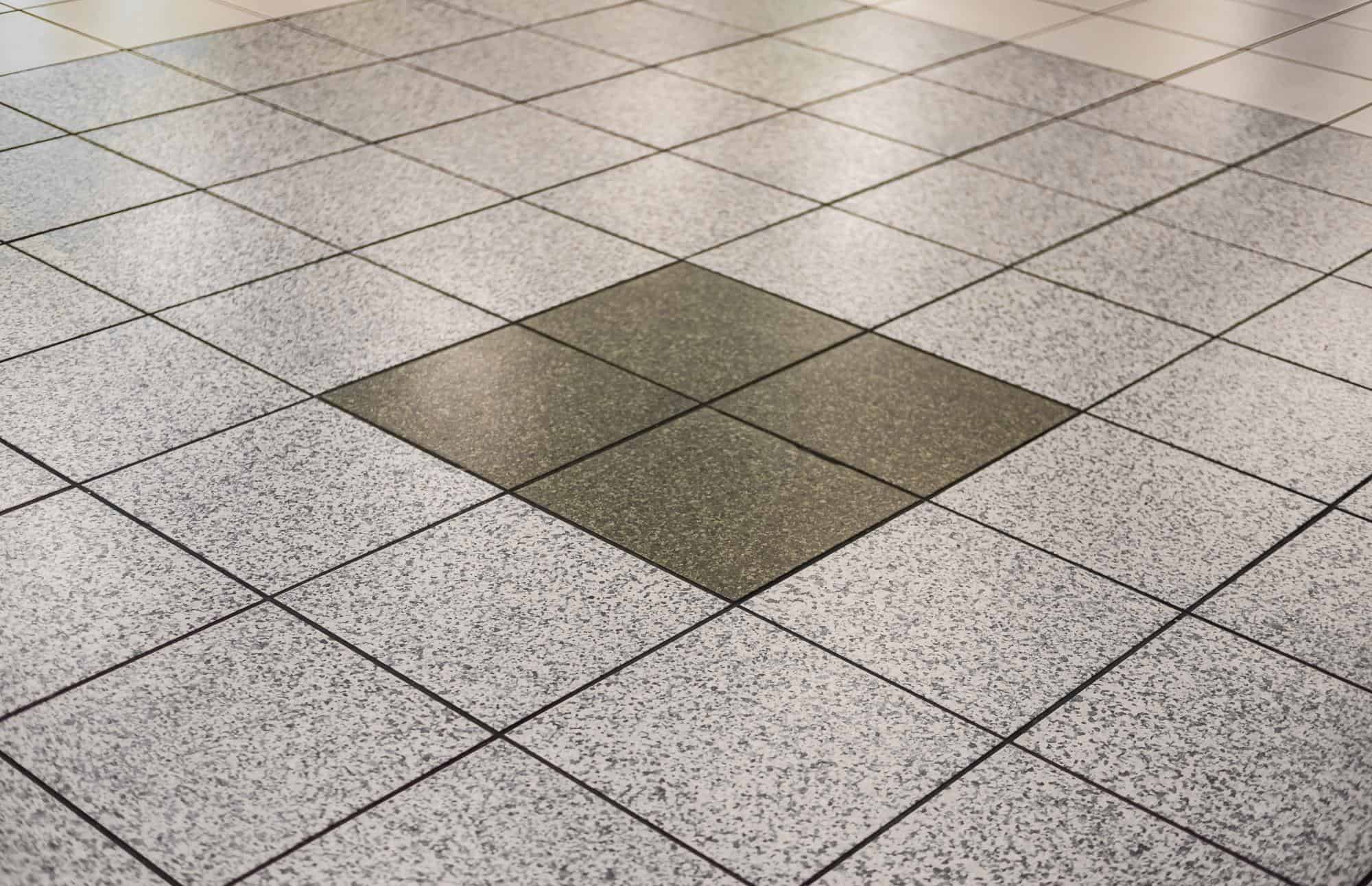 A tiled floor with mostly light gray speckled tiles, featuring a square section of four darker tiles arranged in the center.