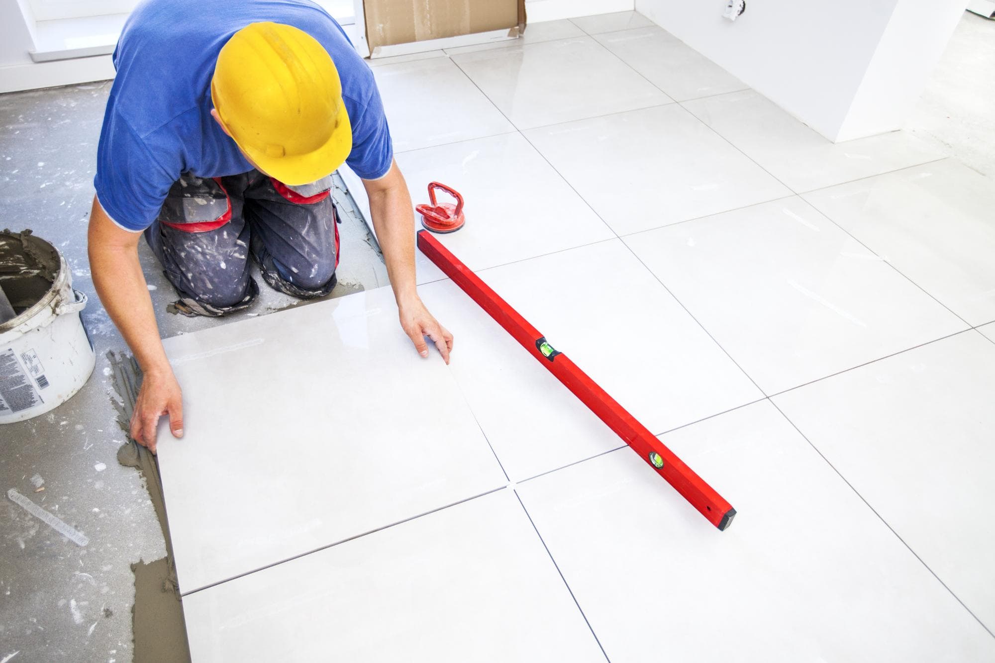 A person wearing a yellow hard hat and blue shirt is installing large white floor tiles. A red spirit level and a bucket of adhesive are nearby, and the person is adjusting a tile for alignment.