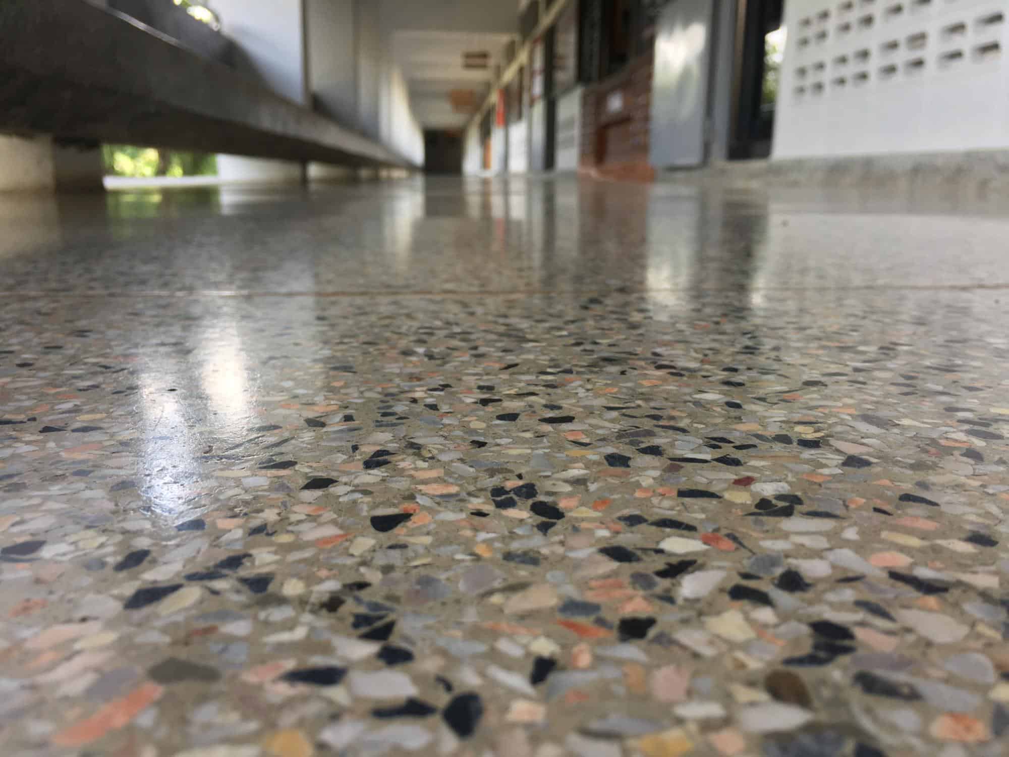 Close-up view of a terrazzo hallway floor in a building, with lockers and a bench visible in the blurred background along the corridor walls.